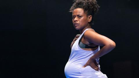 Ayesha Antoine in a Clean Break production called BLANK at the Donmar Warehouse. She has black curling hair pushed up at the top of her head in a loose afro style and short at the back. She is wearing a vest over a bump to suggest she is pregnant