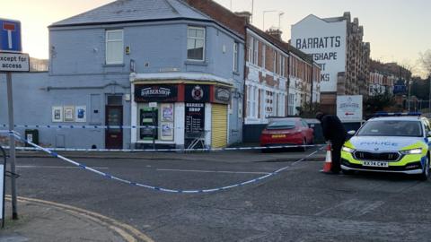 Police tape blocks off a road. A police car can be seen parked on the outside of the cordon while an officer places a cone on the road.