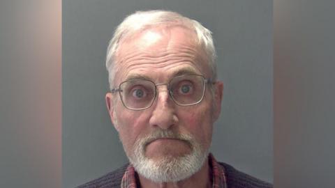 Martin Daniels is wearing glasses, and has grey/white hair and a grey/white beard and moustache. He is wearing a red tartan shirt and a ribbed brown jersey.