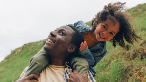 A young girl smiles as she sits on a man's shoulders in the countryside. Both are grinning.