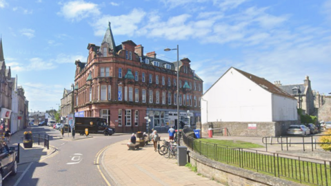 A tall sandstone building stands at the end of a street fillwed with people and vehicles. Blue skies fill the background with a few white clouds.