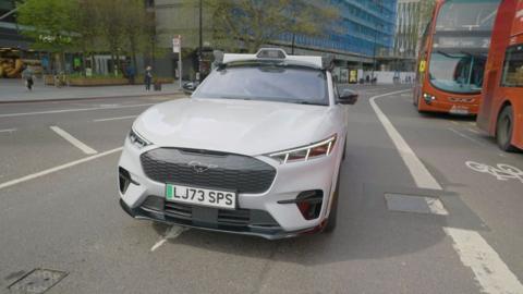 A driverless car on a road in London - there are two red buses to the right