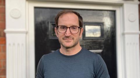 Brendan Mitchell wearing glasses and a blue top stood in front of a black front door