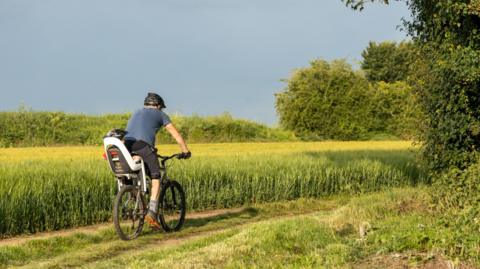 A cyclist pictured from behind on a grassy footpath on a sunny day in the countryside, bordering a field growing wheat. The male cyclist is wearing a grey top, black shorts and black helmet. A child sits in a white seat on the back of the bike, wearing a black helmet. The child's face is not visible.
