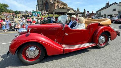 A red classic car with a man and a woman sat inside, is seen at a junction with a church on a raised up area in the distance.