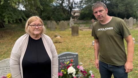 A woman with greying blonde hair and a man stand next to a gravestone decorated with a bouquet of flowers. The man is wearing a green t-shirt and the woman has a black top and a beige cardigan.