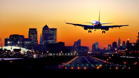 Airplane landing in London City Airport with row of skyscrapers of Canary Wharf and City of London financial districts at background, seen at sunset.