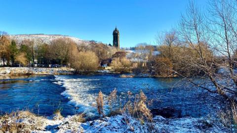 wintry scene showing snow topped hill in distance and river bank, wide view of river, trees with no leaves, tall clock in distance under a blue sky