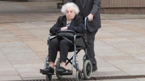 June Mills, with white hair and wearing a black blazer and black trousers, sits in a wheelchair and clutches her handbag as she is pushed away from Liverpool Crown Court by her husband 