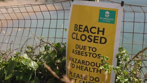 An empty Mount Batten beach near Plymouth which has been closed following the discovery of asbestos. There is a yellow warning signing saying "BEACH CLOSED". The sign is attached to a fence and the sea and beach is blurred in the background.