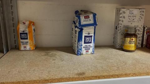 A large shelf showing it looking very bare other than a few bags of sugar and a jar of coffee.