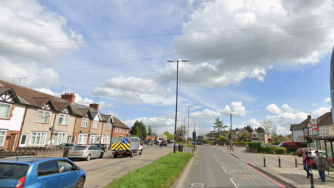 A wide residential road with houses on both sides, parked cars, grassy verges, and a bus stop under a cloudy sky.
