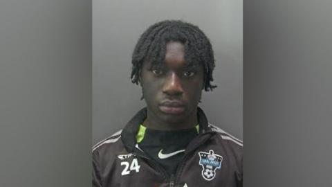 Dodou Manneh is looking at the camera in a police custody shot. He has short black hair in dreadlocks. He is wearing a black jacket and a black T-shirt.