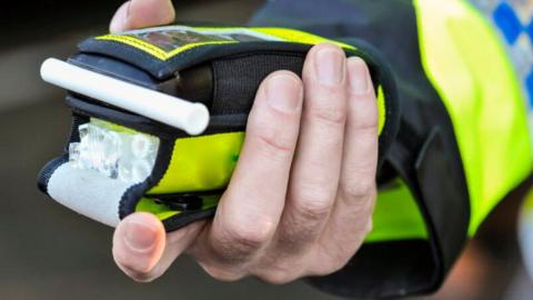 A generic image of a police officer holding a breathalyser in their hand. The officer is wearing a green hi-vis jacket with blue trim. 