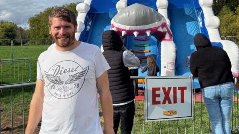 Angus, left, wearing a white T-shirt, standing in front of a large blue inflatable ride with people watching the children using the ride behind him