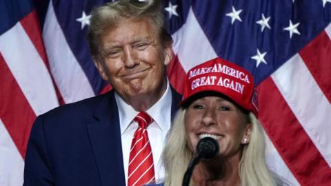Congresswoman Marjorie Taylor Greene, wearing a red cap and speaking into a microphone speaks in support of Trump at his campaign rally in Rome, Georgia in March 2024. Trump looks on in the background, smiling.