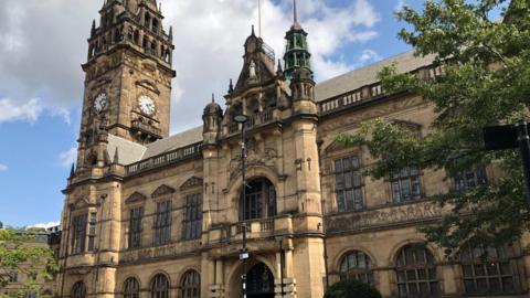 A very grand looking Sheffield Town Hall, complete with the town hall clock reading 2.25pm.  A sandstone Victorian building pictured on a sunny day, with a mostly blue sky background and two trees in full bloom in the foreground on the left and right of the building.