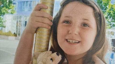 Minnie-Rae Dunn smiles at the camera. She had brown hair held back by a band and carries a small teddy bear.