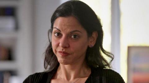 A woman with dark medium length hair and brown eyes is looking at the camera. She is wearing a black cardigan and a chain with a small pendant around her neck. The background is blurred, but shows a bookcase and light coming through a window.