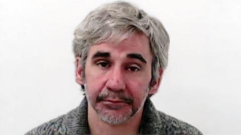 A mugshot of a man with long grey hair and dark eyebrows. He is wearing a grey knitted top.