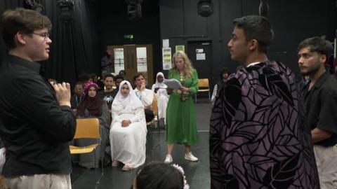 A group of teenage students rehearsing a scene from a Shakespeare play. There are two boys standing in the foreground, one wearing a cape, and several people sitting down watching. A woman in a green dress holds a script watching them. 