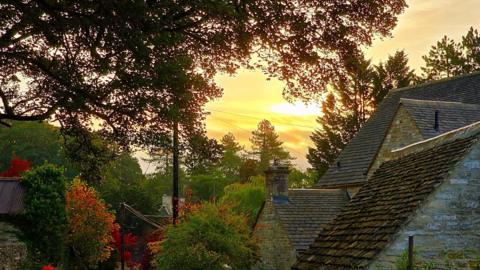 Sun setting behind a thatched cottage with autumnal trees in the foreground 