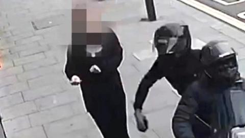 screengrab of security camera where two people in helmets on a moped are taking phone from a woman's hand