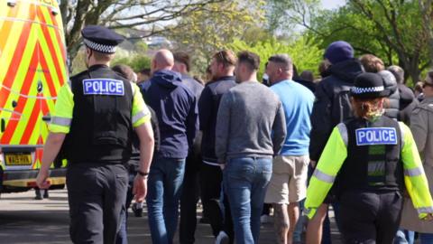 Two Devon and Cornwall Police officers walking with a number of football supporters outside Home Park Stadium in Plymouth. They all have their backs to the camera. There is a police van parked in the distance along with a number of trees.