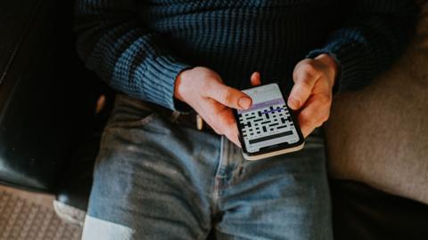 A man, wearing blue jeans and a dark blue shirt, at home holds a smart phone and uses a cryptic crossword app.