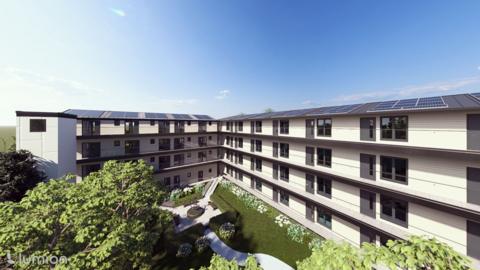 An illustrative image of the new hospital. It is a four-story white building with rows of windows on each floor. The roof is mostly flat with solar panels on top. There is a green space outside with grass, flowers, trees and a seating area.