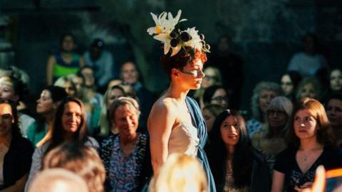 A woman walking on a catwalk wearing a white and blue dress and white lilies on her head. People are watching from the sides.