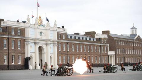 Troops fire a gun in front of the army barracks at Woolwich, an elegant brick structure with a white painted arch mounted by flags and a Royal coat of arms. The flash of the gun is visible in the centre of the image. 