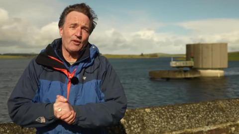 David Braine standing next to a reservoir. He is in a blue jacket looking at the camera. His hands are held together. It is a cloudy day.