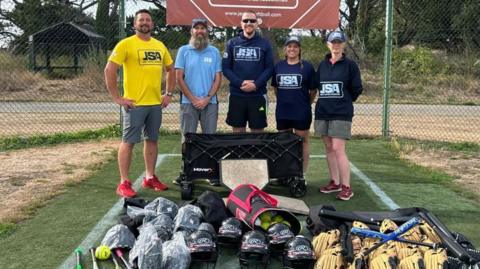 Three men and two women from the Jersey Softball Association stand behind a load of softball equipment. There are stood in a batting cage with Jersey Softball Association banners of the fencing. 