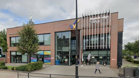 Street view image of the college on a cloudy day. It is a rectangular shape with two storeys and has large glass windows at the front. People are walking past the building and there is a tree with some greenery to the side of it. 