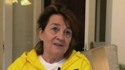 A woman with shoulder length brown hair is wearing a yellow zip-up hoodie over a white T-shirt