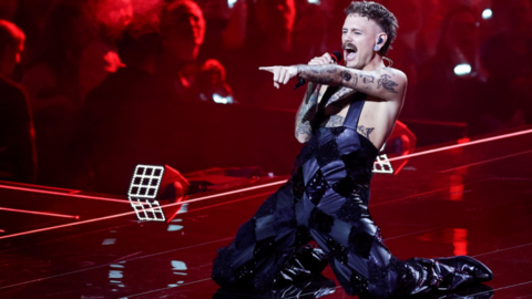 A performer is captured mid‑emotion onstage, kneeling with one knee down and the other bent, body leaning forward into the microphone. They’re dressed in a shiny, patterned sleeveless outfit that reflects the red stage lights, and their tattoos stand out across their arms and torso. The lighting is intense and saturated — deep reds flooding the stage — giving the whole moment a sense of heat, urgency, and raw energy. Behind them, the crowd glows in scattered points of light, phones raised. Their posture — head tilted, mouth open mid‑note, hand gripping the mic