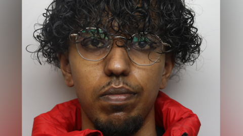 A police mugshot of a man with curly black hair, glasses and a red coat.