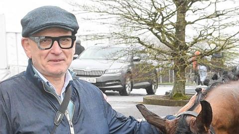 A man in a flat cap smiles as he holds a racehorse. He is wearing thick rimmed black glasses, a blue shirt and navy blue jacket. The horse is being held by its reins and is bowing its head. The are standing in front of a car park, a sole car can be seen in the background, parked next to a tree.