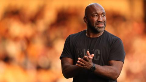 Port Vale manager Darren Moore in a black club t-shirt claps as he leaves the field