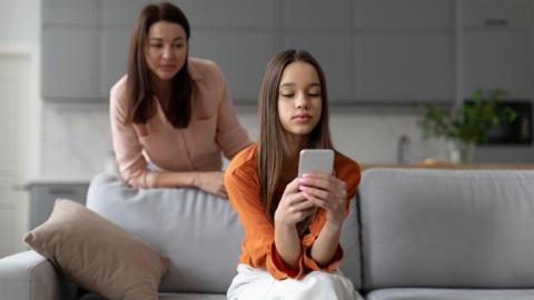 A teenage girl looks at her phone with her mother watching over her shoulder