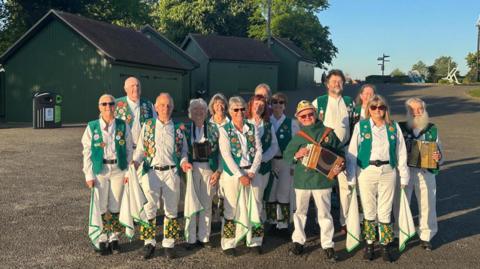 A group of Morris dancers are gathered in Promenade Park, Maldon. They are wearing traditional costumes with green waist coats. The group are smiling and some members are holding instruments. They are holding scarves to use for dancing.