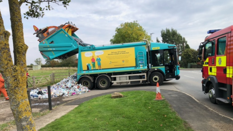 Blue refuse lorry with the back open after dumping plastic waste in a layby. There is a fire engine. There are trees and grass.