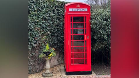 A red phone box standing in the corner of a garden against an ivy clad wall. The phone box looks in pristine condition after being refurbished. 