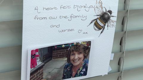A piece of white paper stuck to a window which has an image of a woman with brown hair smiling on it. Also on the paper is a drawing of a bee and a message which reads 'A heartfelt thank you from all the family and Worzel'