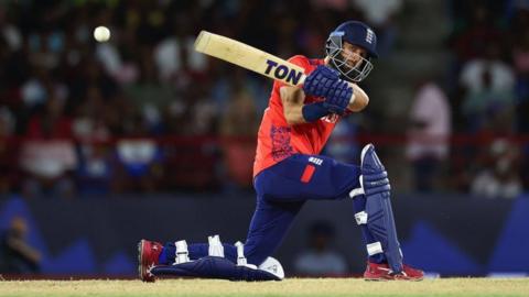 Moeen Ali is in navy and red cricket clothes, with the England logo on them. He has a helmet on, and is holding a cricket bat, taking a swing. He is also wearing navy gloves and navy shin pads.