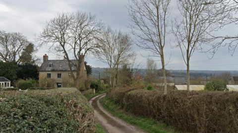A narrow country lane with tall hedges either side of it runs past a large three-storey old house.