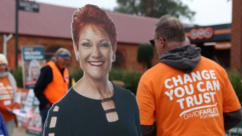 A cardboard cutout of Pauline Hanson on the left with a man in an orange campaign t-shirt on the right in front of a polling station.