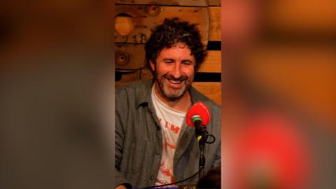 Mark Watson smiles as he talks into an orange microphone.