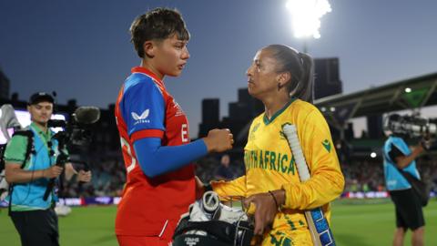 Issy Wong (left) of England and Shabnim Ismail (right) of South Africa interact following the ICC Women's T20 World Cup 2026 semi-final at the Oval. The taller Wong wears red cricket gear with blue arms while Ismail is all in yellow with the words 'South Africa' written in green across her chest. She also has a bat handle tucked under her left arm, which is nearest the camera, and clutches a helmet with batting gloves in it. Stewards in blue bibs move around them while a floodlight in the darkening sky shines down brightly above a stand